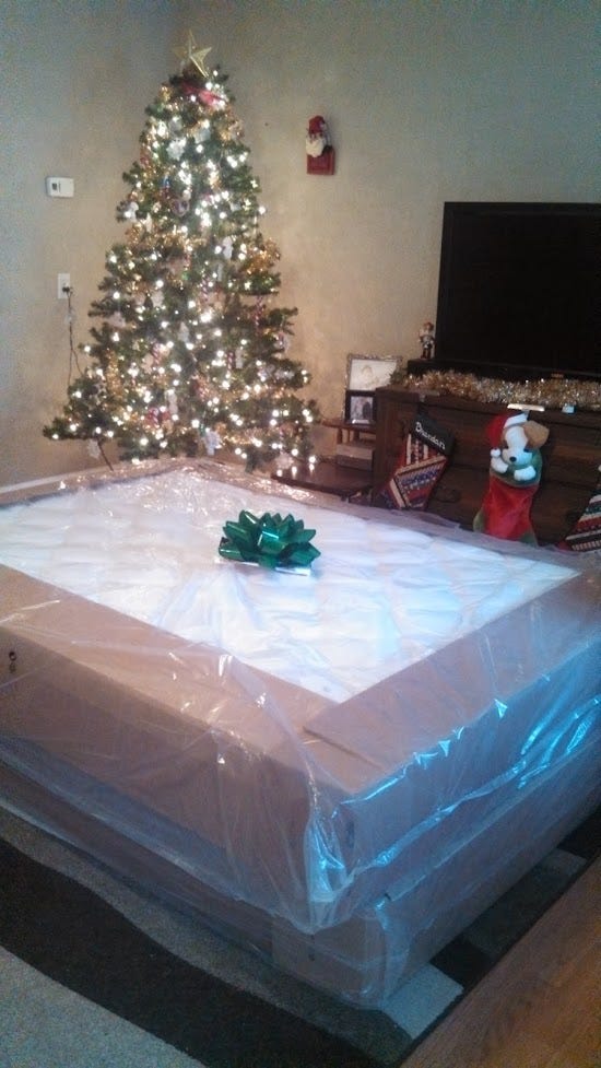 New mattress and boxspring still in plastic with a giant green gift-bow on top, in front of a Christmas tree and stockings hanging from the entertainment center.