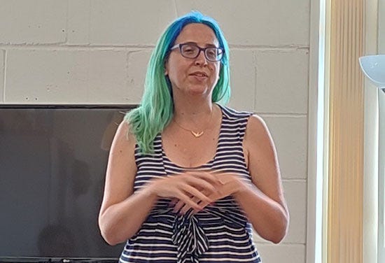 Alice, a woman with bright blue and teal hair, stands to talk to the room.