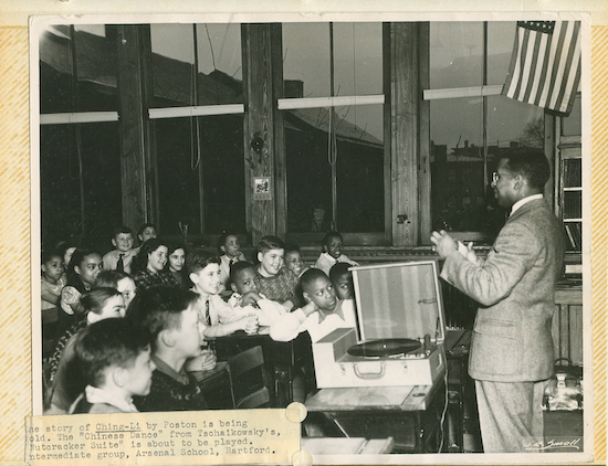 historical photo of a black male librarian in a suit talking to a classroom if kids of various races. He is standing by a record player. A flag with 48 stars hangs over him.