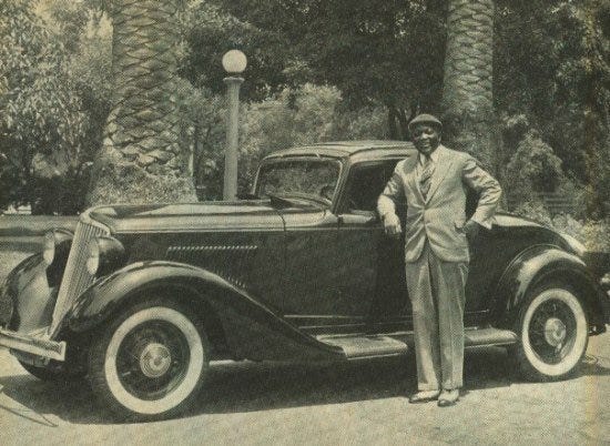 A black and white photograph of a man in a suit standing next to an expensive car
