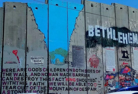 Photo of a portion of gray concrete wall covered in graffiti. The word Bethlehem is painted in all caps, and a huge fissure is painted on the wall. There is a large block of text reading, “ ‘Here are God’s children on both sides of the wall, and no man made barrier can destroy this fact,’ ” King shouted. ‘With this faith we will be able to tear out of the mountain of despair a stone of hope.”