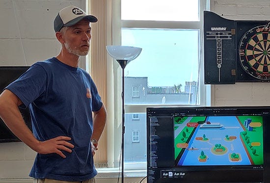 Stuart, a man in a baseball hat and blue t-shirt, stands next to a TV showing a cartoony illustration of a cruise ship and small islands in a river
