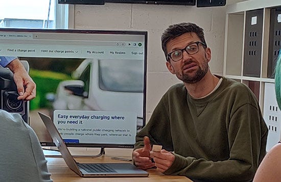 Neal, a man in a green sweatshirt and black rimmed glasses, sits next to a monitor showing the website for Chargy.
