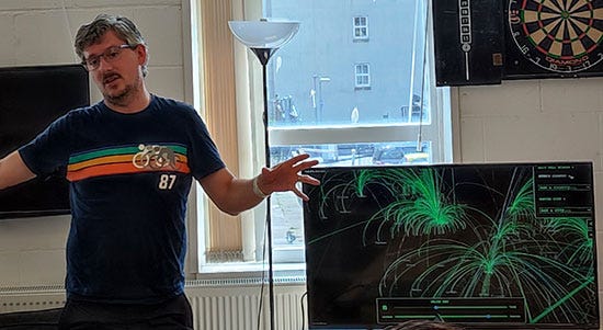 Tom, a man with glasses and a black t-shirt with a rainbow on, stands next to a monitor showing a map with arcing lines, rather like you’d see in a film about nuclear war showing the worst case scenario.