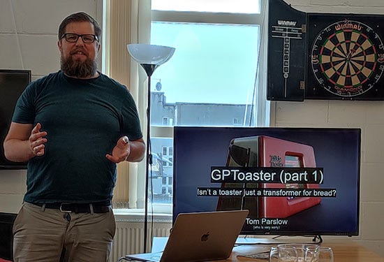 Tom, a man with glasses and a full beard, presents a talk on using a large language model to mimic Talkie Toaster from the TV show Red Dwarf