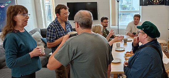 In the foreground four people stand drinking coffee and chatting, in the background more are sitting around a long kitchen table and chatting with pastries and drinks