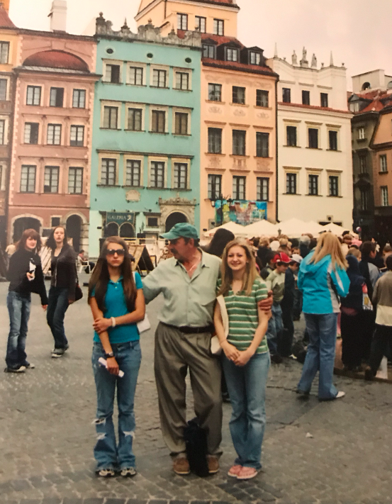 A man with his arms around his two teenage daughters on a Polish street. One is smiling and the other looks embarrassed.