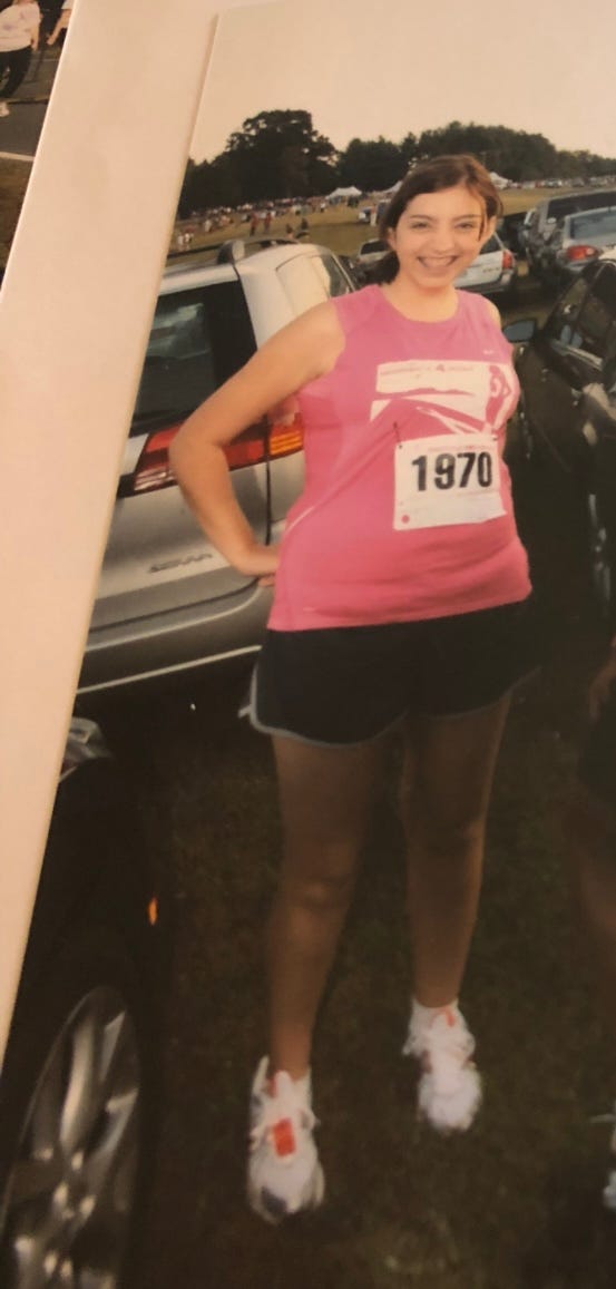 A white female teenager of around 16 smiles at the camera. She has braces and her hair is pulled back in a ponytail. She is wearing a tank top with the number “1970” on it and short black shorts. She is outside and several cars are visible in the background.