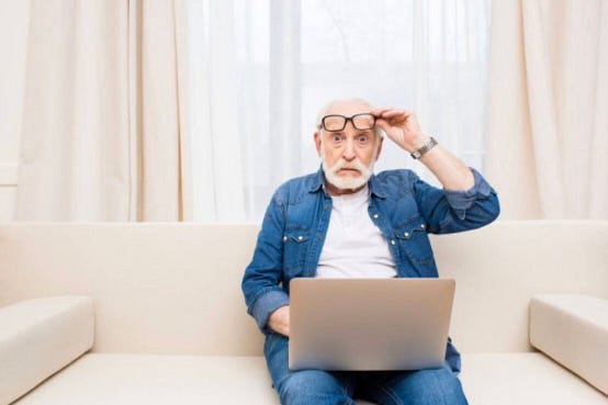 #pratodosverem: um idoso usando roupa jeans e um óculos de grau preto, sentado em um sofá, com o fundo bege claro. Ele está com uma expressão assustada segurando e olhando um notebook.