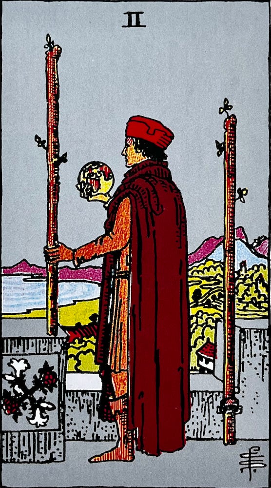The Two of Wands — A man stands on a balcony looking out over a shoreline scene. He holds a golbe in his right hand and a wooden staff or wand in his left hand.