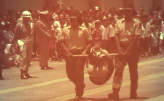 Dois indígenas, com uniformes da GRIN, carregam uma pessoa em um pau de arara em demonstração na formatura.
