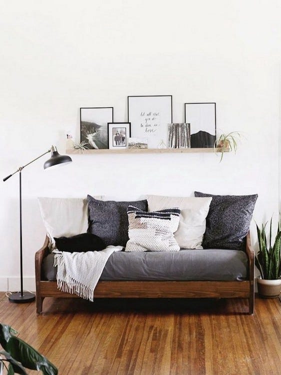 A loveseat with gray cushions and decorative pillows on a wood frame under open shelving with art prints and house plants.