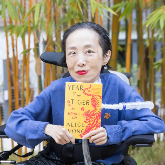 Photo of Alice Wong, an Asian American disabled woman in a power chair, against a background of bamboo trees. She is wearing a blue cardigan and sitting in a power chair. She is holding a copy of her memoir, Year of the Tiger, a paperback in yellow and red with a fierce tiger on it designed by Madeline Partner. She is wearing a bold red lip color and a trach at her neck. Photo credit: Eddie Hernandez Photography.