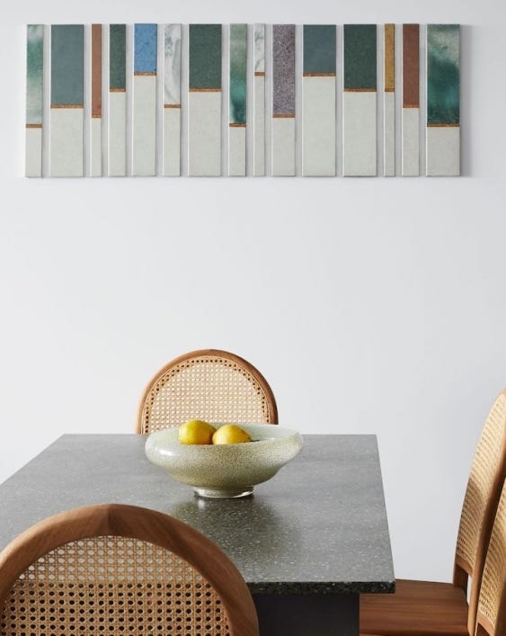 A photograph of a dining room. There is a grey dining table, with a bowl with two lemons on it and two chairs with brown net backs. Above the table on the wall is a role of narrow tiles as wall art. Each has a white bottom section and a coloured and textured top section at varying heights.