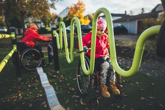 Duas crianças de pele branca, em cadeiras de roda, vestindo roupas de inverno. Brincam em um parque projetado com design inclusivo.