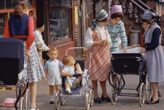 A group of Satmar women on a city street, chatting and walking strollers. They wear kerchiefs over their wigs.