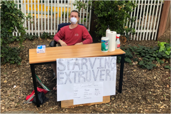A young man sitting at a desk with a sign that reads, “Starving Extrovert.”