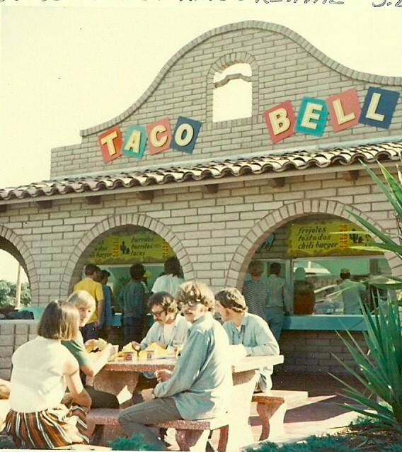Slightly sepia-toned picture of a group of people eating at an outdoor table in front of an old-school Taco Bell.