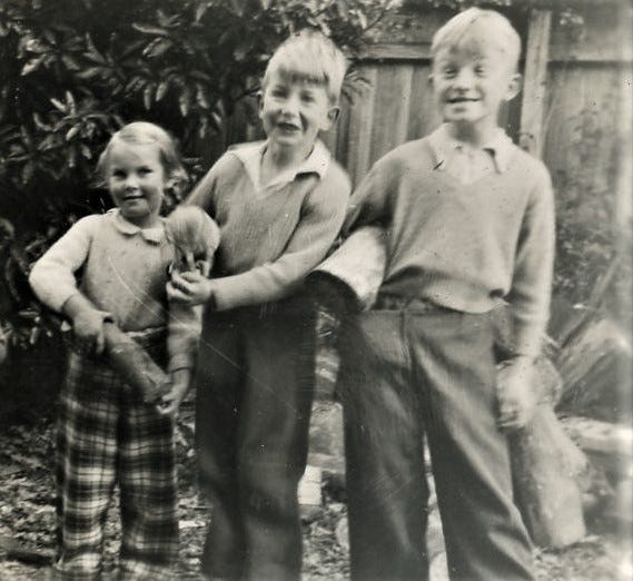 A black and white picture from the 1950s. Three young children stand together in a backyard in front of a pile of firewood. All smile; the middle one holds a duckling pecking feed out of his hand; the boy on the right, taller than the others, holds a wooden log and is in the middle of making a goofy face.