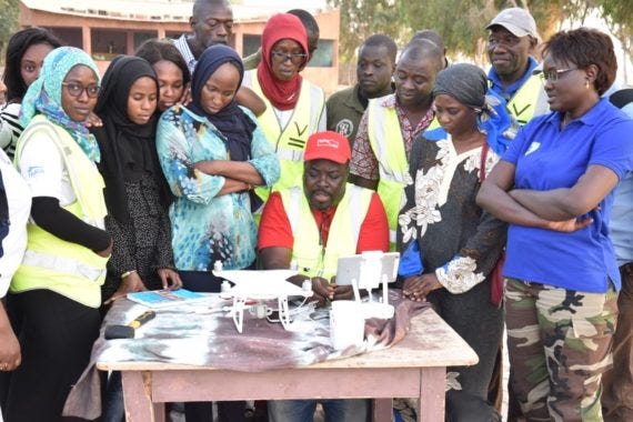 A group of community members surround a man in a red cap giving a drone demonstration on a table. Many are wearing highlighter yellow construction vests.