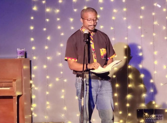 Photo of Professor Dr. Doug Manuel reading from his newest poetry collection “Trouble Funk” in front of lights.