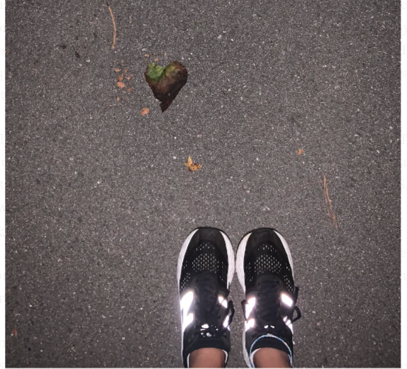 Image shows Candace’s black sneakers on a paved running trail. There is a heart-shaped leaf to the left of where she stands.