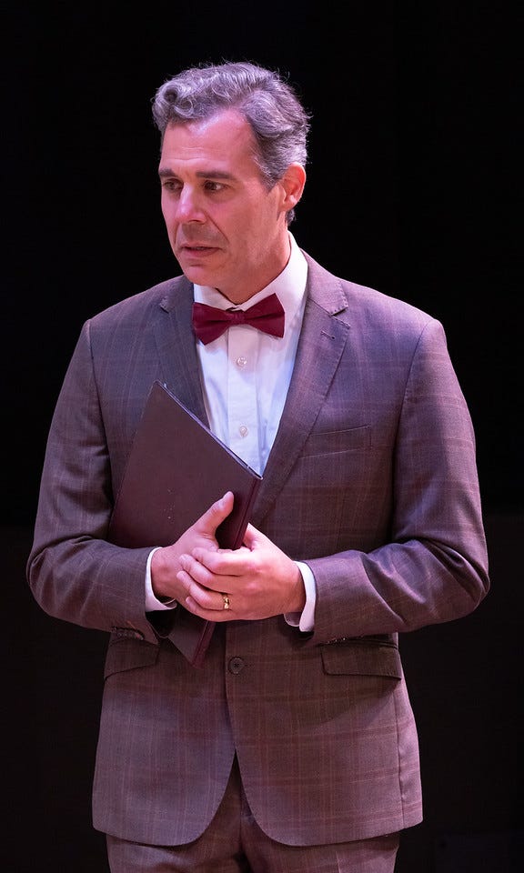 A man with olive skin and short gray hair in a gray suit holds a folder, looking nervous.