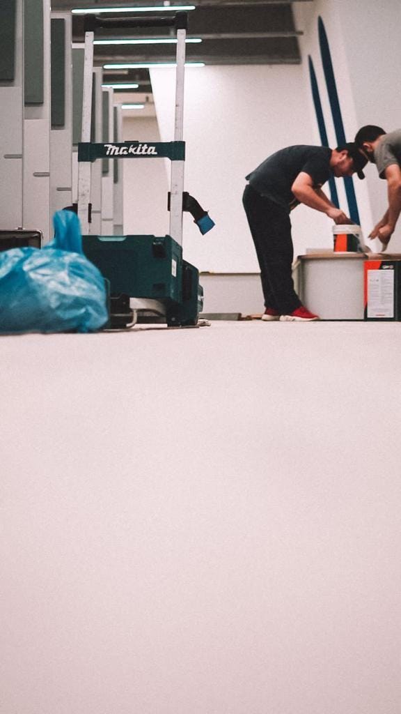 Foto de um cantinho na área de estações de trabalho sendo construído. Há apenas paredes e um banco branco, e nota-se dois homens à direita abrindo um balde que parece ser de algum material de construção.