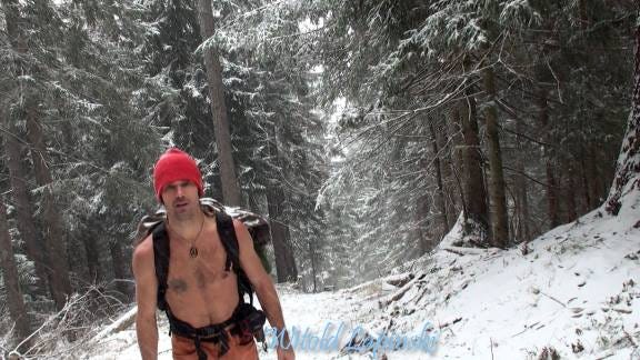 A man with naked upper body and a red bonnet walks through a forest with snow.