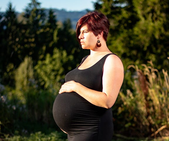 Short-haired pregnant woman with trees and sky behind her