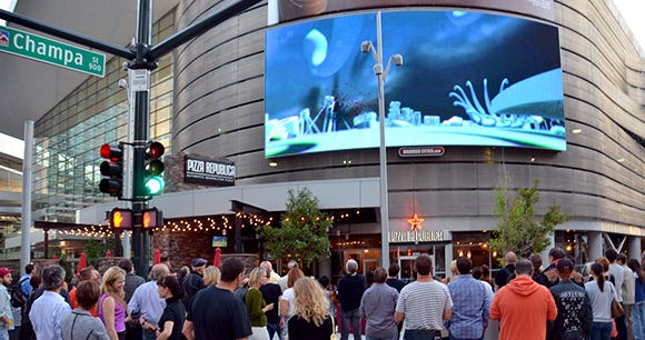 A crowd of people outside the Denver Convention Center watching an experimental short film being played on a large LED billboard.