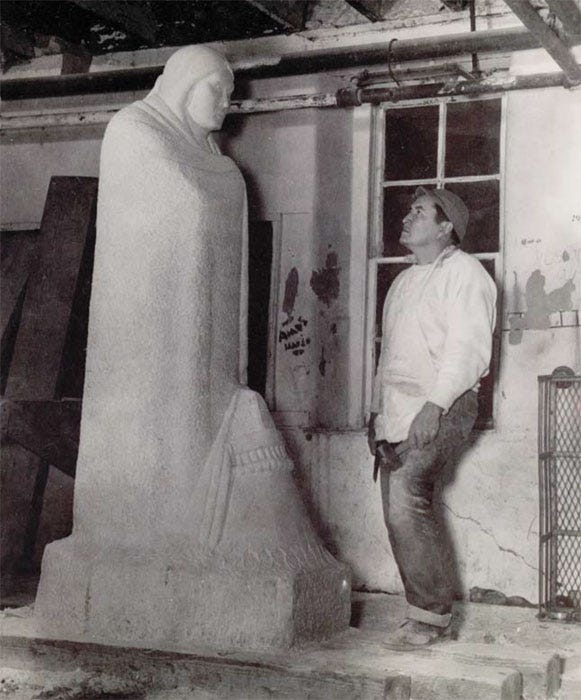 Sculptor standing with tool in hand, looking up at the face of his sculpture, a solemn Native American figure.