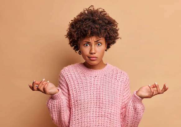 Mulher jovem, preta, cabelo black power, usa uma blusa de manga comprida na cor rosa e na sua expressão aparenta estar confusa sobre algo.