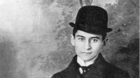 A imagem mostra Kafka jovem usando um chapéu preto contra um fundo acinzentado. Ele usa um terno preto e olha em direção da câmera com um sorriso.