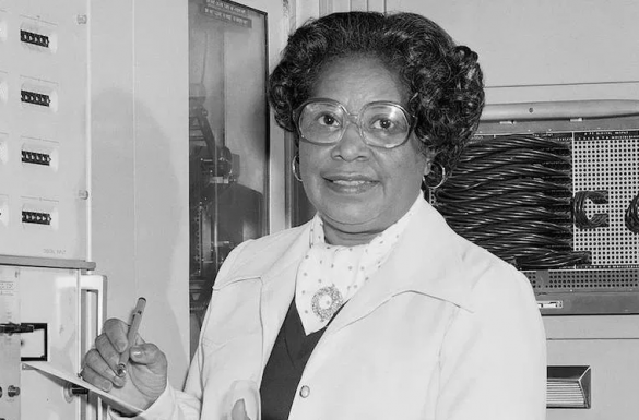 A mid-aged woman standing in front of equipment found in a science lab. She is wearing glasses with a round frame and a white blazer with a dark sweater under it. She is holding a pen and a writing pad.