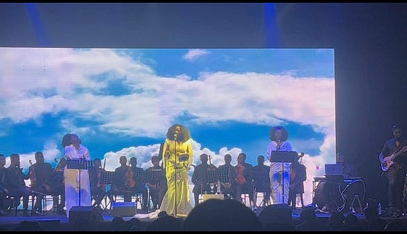 Titilope Sonuga stands centre stage in a white full-length dress flanked on both sides by backup singers from the MelAfrique band wearing white jumpsuits. Behind them sits an ensemble of violinists with a screen of white clouds in a blue sky in the background.