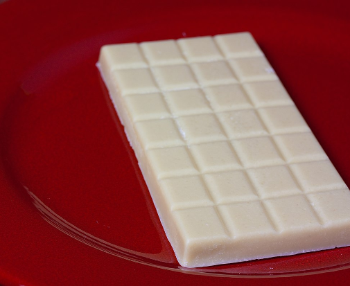 A photograph of a vegan white chocolate, scored in squares bar. It is placed on a red shiny circular surface. The background is blurred.