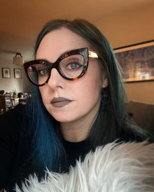 A pale, femme-presenting person with long, dark blue hair, chunky cat-eye glasses, a septum piercing looking unsmiling into the camera