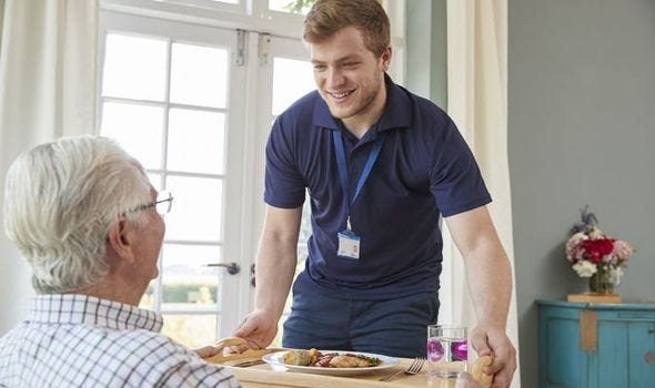 A male care worker brings dinner to older male in care home