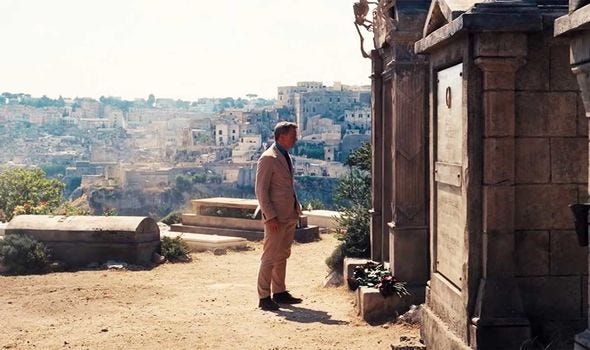 Daniel Craig as James Bond dressed in a tan coloured suit stands opposite a stone Mausoleum high on the hills above the Italian town of Matera which can be seen behind him.