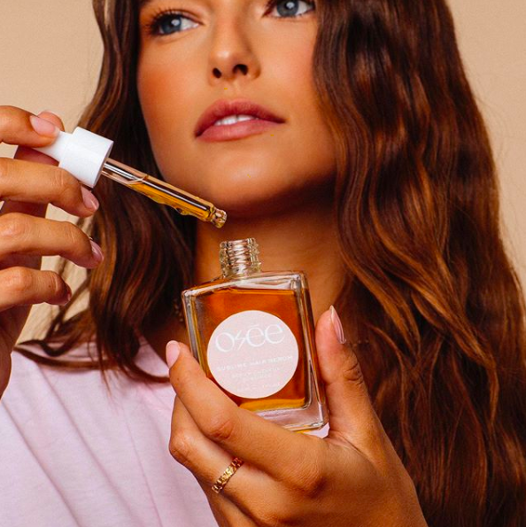 A woman holds a glass bottle and dropper containing Osee hair serum.