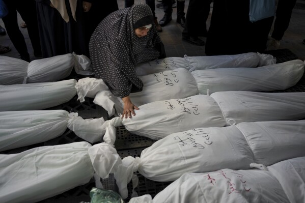 Uma mulher palestina e muçulmana ajoelhada e observando os corpos de palestinos assassinados no genocídio. Eles estão cobertos com panos brancos.