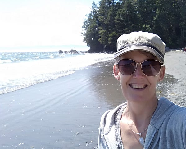 Photo of author, Gill McCulloch standing on a sandy beach in Sooke, BC, Canada. The sun is shining on waves with trees in the background.