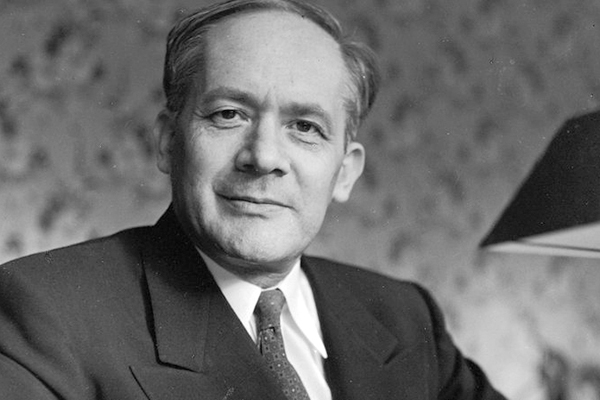 A imagem mostra um Raphael Lemkin de terno, sorrindo. Ele está vestido com roupas formais e está em casa. A foto está em preto e branco. Ele olha em direção para a câmera.