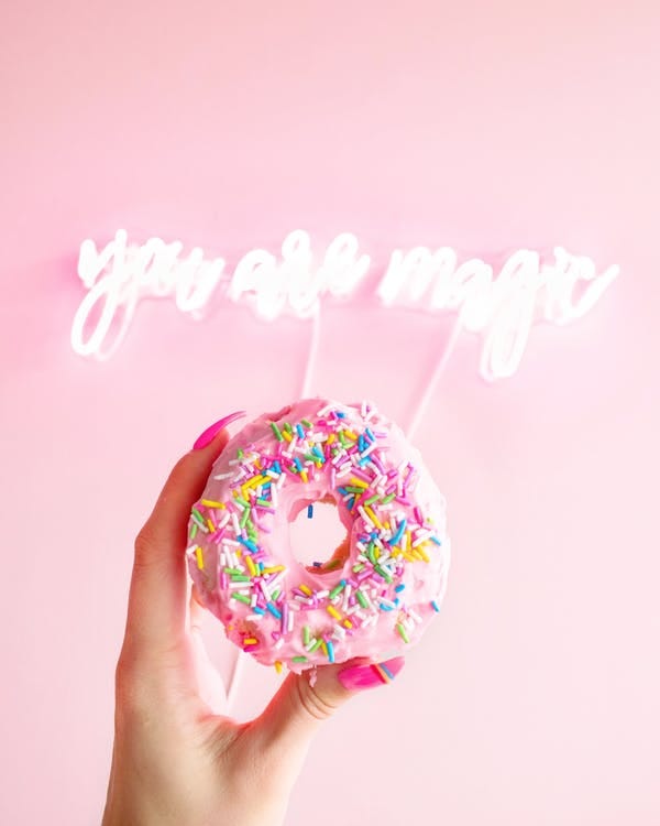 A hand holding a donut with “you are magic” in neon on the wall in the background