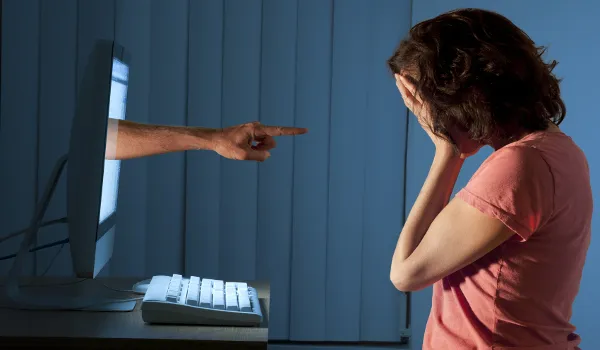 Uma mão saindo da tela de um computador apontando com o dedo indicador de forma incisiva para uma jovem que está com as mãos protegendo o rosto, demonstrando um sentimento de pavor ou desespero.