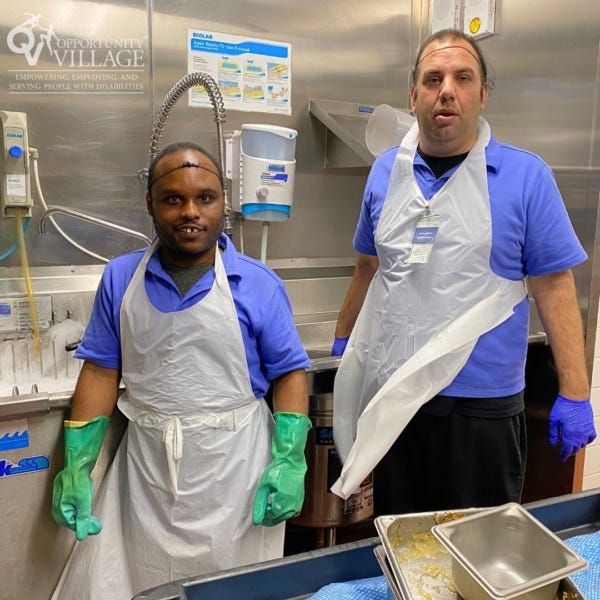 two people working in a commercial kitchen wearing aprons and gloves