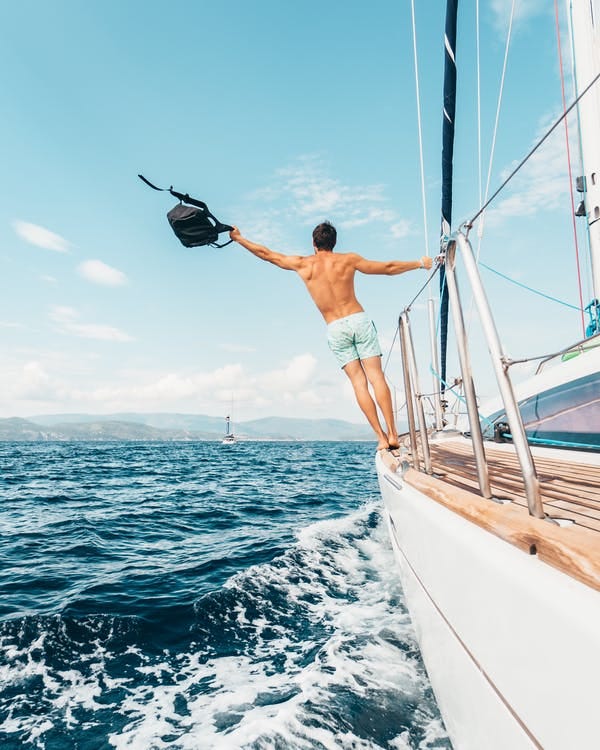 A man with a backpack in his hand hanging off the railing of a sailing yacht with a carefree pose.