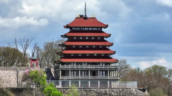 Red multi-tiered pagoda with traditional East Asian architecture, set against lush trees and a…