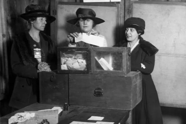Três mulheres em volta da urna com papéis para votação.